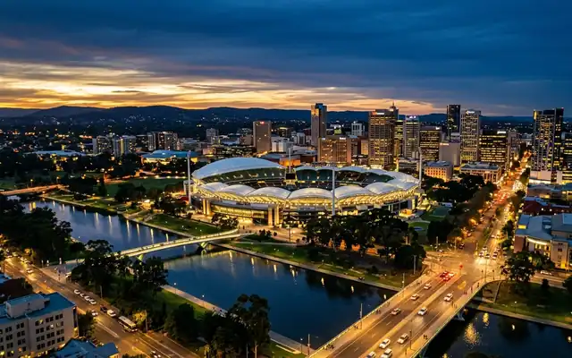Adelaide Oval's scalloped white roofs beside the River Torrens with the city skyline at dusk