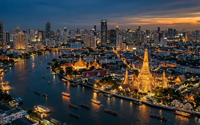 Wat Arun's illuminated spires beside the Chao Phraya River and Bangkok high-rises at dusk
