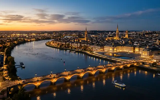 The arched Pont de Pierre over the Garonne with Bordeaux's lit riverfront and church spires at dusk