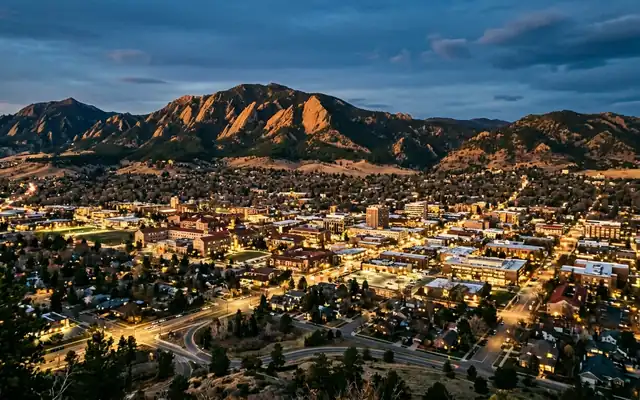 Boulder at dusk, the tilted sandstone slabs of the Flatirons rising behind the low city grid and campus rooftops
