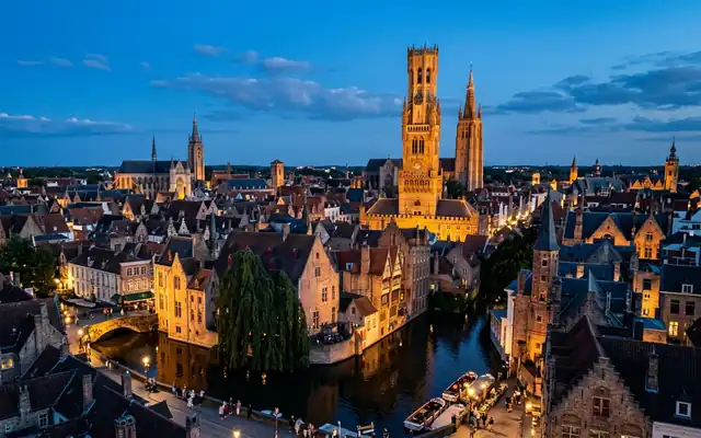 Brugge old town at dusk, the medieval Belfry tower above stepped-gable rooftops and a tree-lined canal