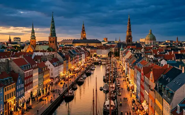 Colourful gabled townhouses and moored boats along Nyhavn canal in Copenhagen with spires beyond at dusk