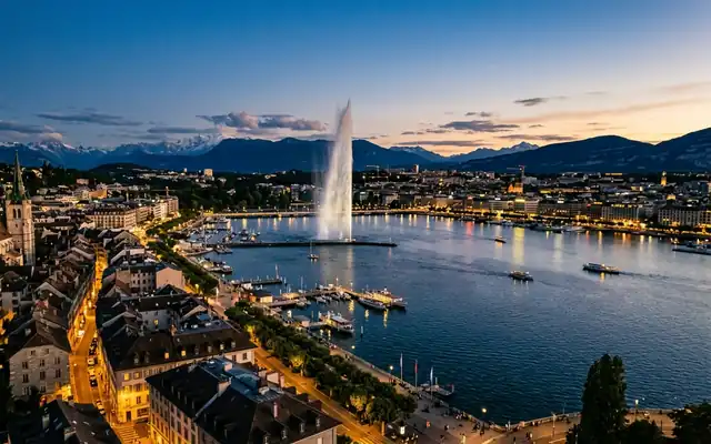 The Jet d'Eau fountain over Lake Geneva with Geneva's lit old town and mountains at dusk