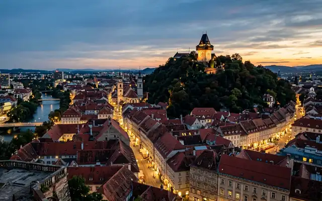 The Schlossberg clock tower above the red-tiled rooftops of Graz's old town at dusk