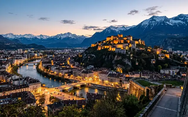 Grenoble at dusk, the hilltop Bastille fort and its cable cars above the Isere river with snow-capped Alpine peaks behind