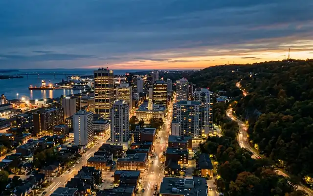Hamilton downtown at dusk, lit office towers between the harbour with its cargo ships and the wooded Niagara Escarpment ridge