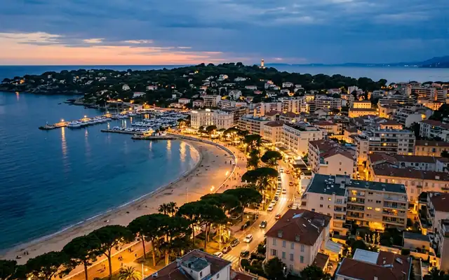 Juan-les-Pins at dusk, the curving sandy bay and pine-lined promenade leading toward the Cap d'Antibes lighthouse headland