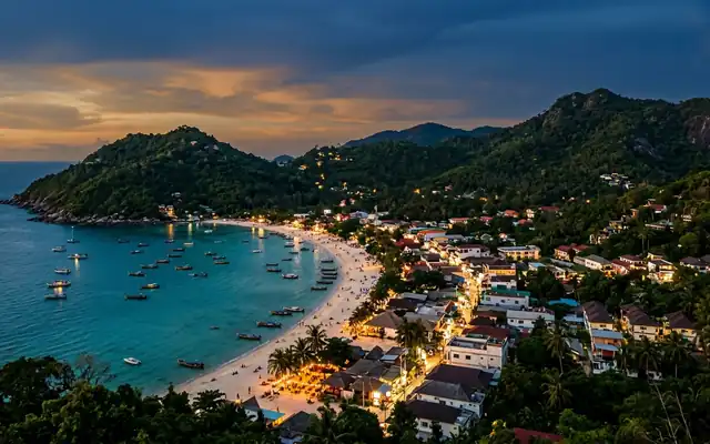 A curving sandy bay on Koh Phangan with longtail boats and beachfront village lights at dusk