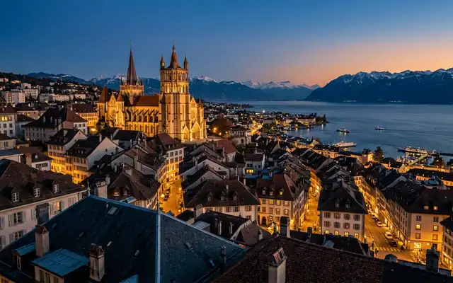 Lausanne old town at dusk, the Gothic cathedral above tiled rooftops with Lake Geneva and the snowy Alps beyond
