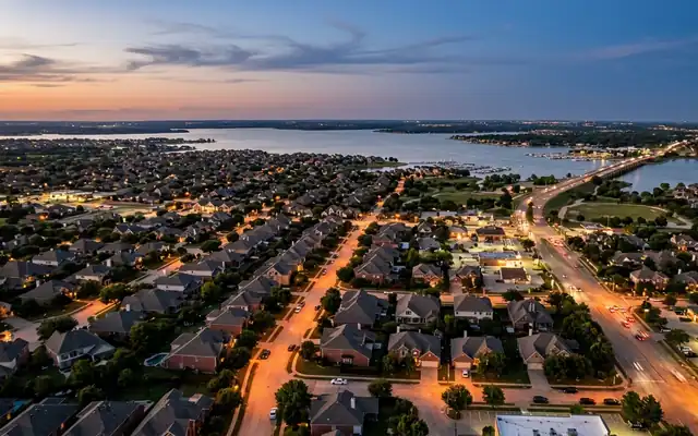Lewisville at dusk from above, North Texas suburban rooftops and lit streets reaching the wide shoreline of Lewisville Lake