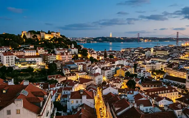 Lisbon's terracotta rooftops below Sao Jorge Castle, with the Tagus and 25 de Abril bridge at dusk
