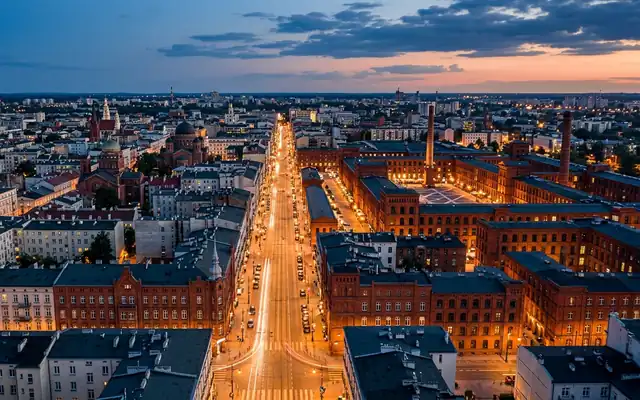 A long straight lit street through Lodz flanked by red-brick factory buildings at dusk