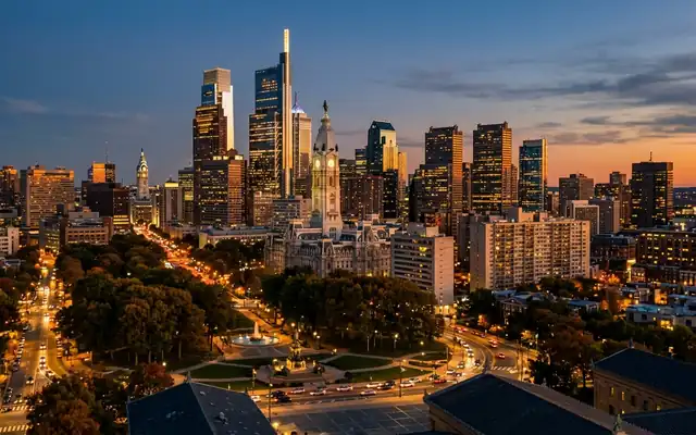 Philadelphia City Hall's clock tower amid the Center City skyline above a tree-lined parkway at dusk