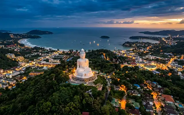 The white Big Buddha statue on its hilltop above Phuket's lit beach bays at dusk