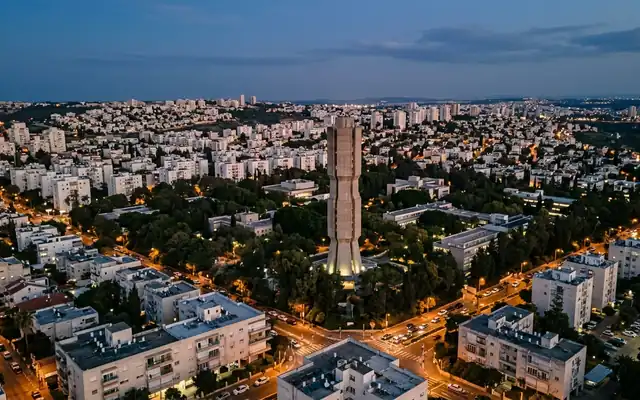 Rehovot at dusk, the ribbed concrete Koffler accelerator tower rising over the leafy Weizmann Institute campus and white apartment blocks