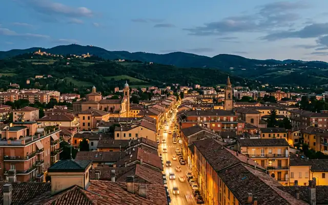San Lazzaro di Savena at dusk, terracotta rooftops along the lit Via Emilia with green Apennine foothills behind