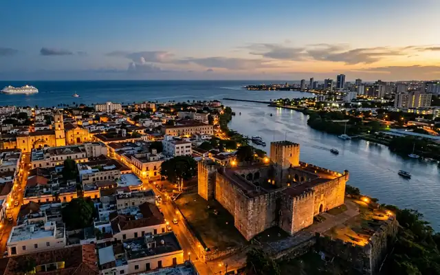 A stone fortress at the river mouth with the colonial rooftops of Santo Domingo at dusk