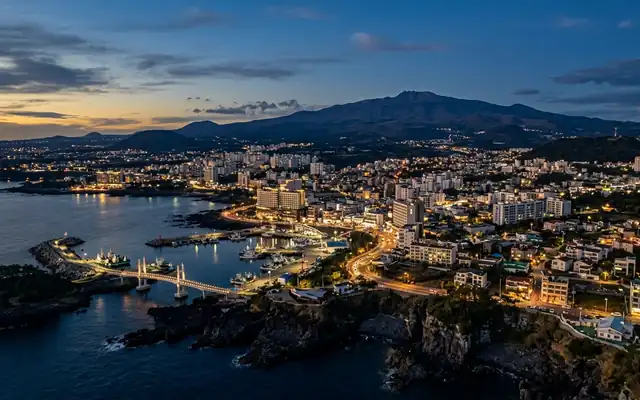 Seogwipo on Jeju at dusk, the harbour town on its volcanic shore with the broad shield of Hallasan behind