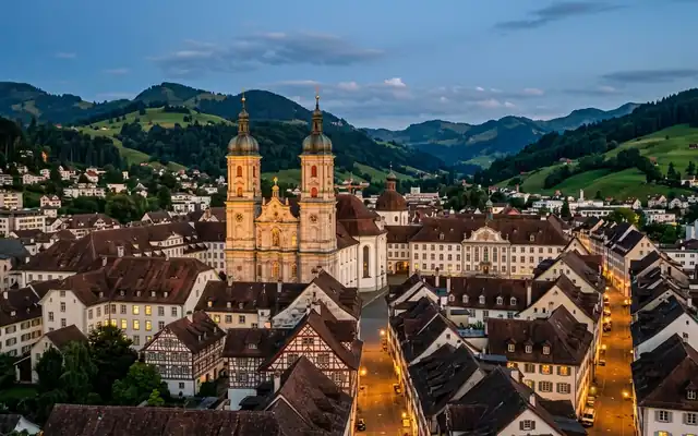 St. Gallen old town at dusk, the twin baroque towers of the Abbey Cathedral above steep tiled rooftops and green Appenzell hills
