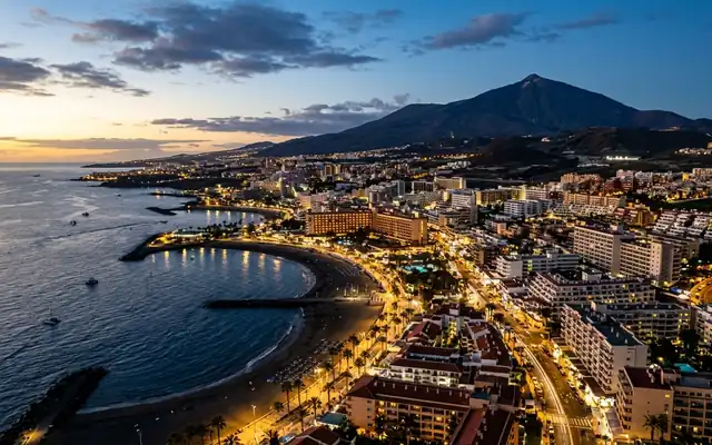 The lit beachfront of Playa de las Américas in Tenerife with Mount Teide rising behind at dusk