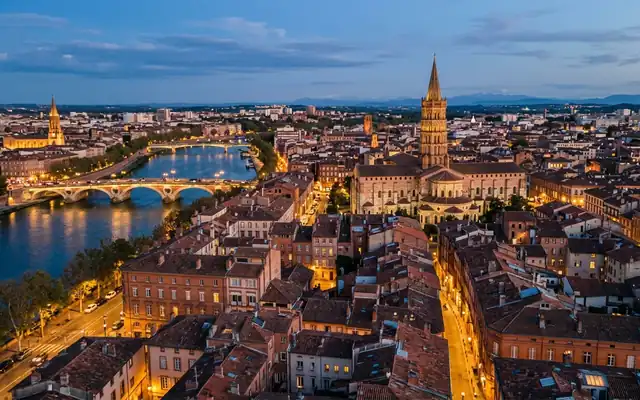 Toulouse at dusk, the octagonal brick bell tower of Basilique Saint-Sernin above pink rooftops with the Pont Neuf crossing the Garonne