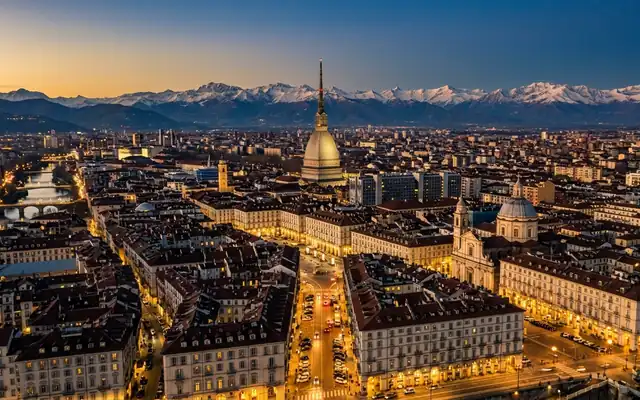 Torino at dusk, the Mole Antonelliana spire above the grid of rooftops with the snow-capped Alps on the horizon