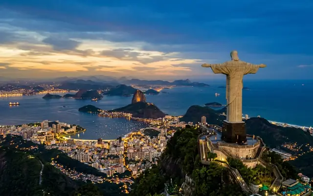 Christ the Redeemer above Rio de Janeiro, Brazil at dusk, arms outstretched over Sugarloaf Mountain and the lights of Guanabara Bay