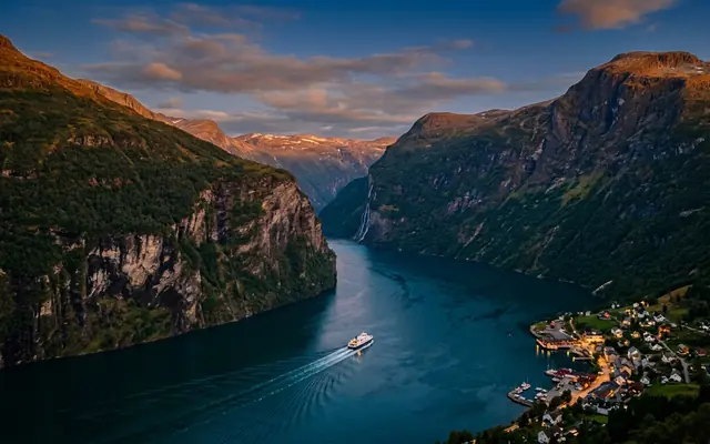 Geirangerfjord in Norway at dusk, a small ferry tracing a wake between steep fjord walls, a lit village on the shore