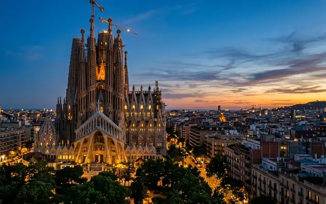 The Sagrada Familia basilica rising over Barcelona at dusk, its Gaudi spires and construction cranes above the surrounding rooftops