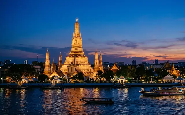 Wat Arun lit at twilight on the Chao Phraya River in Bangkok, its tiered prang spire above boats on the water