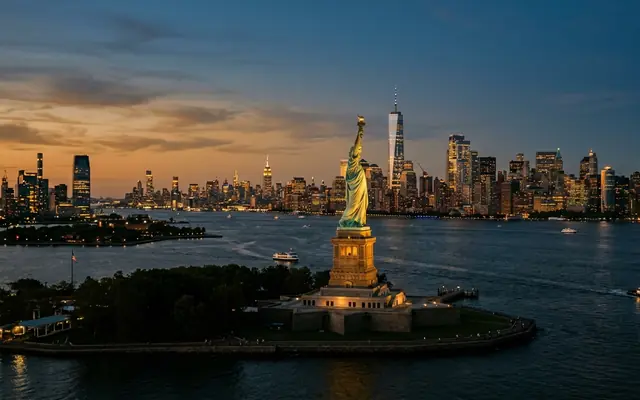 The Statue of Liberty on Liberty Island at dusk, torch raised against the lit Manhattan skyline across New York Harbor