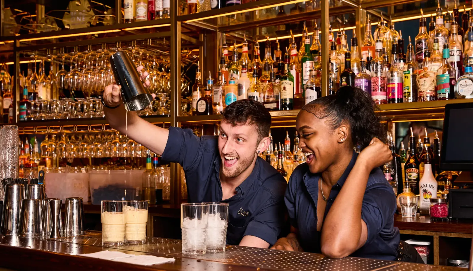Bartenders mixing cocktails behind the bar at Be At One Liverpool Street with shelves of spirits