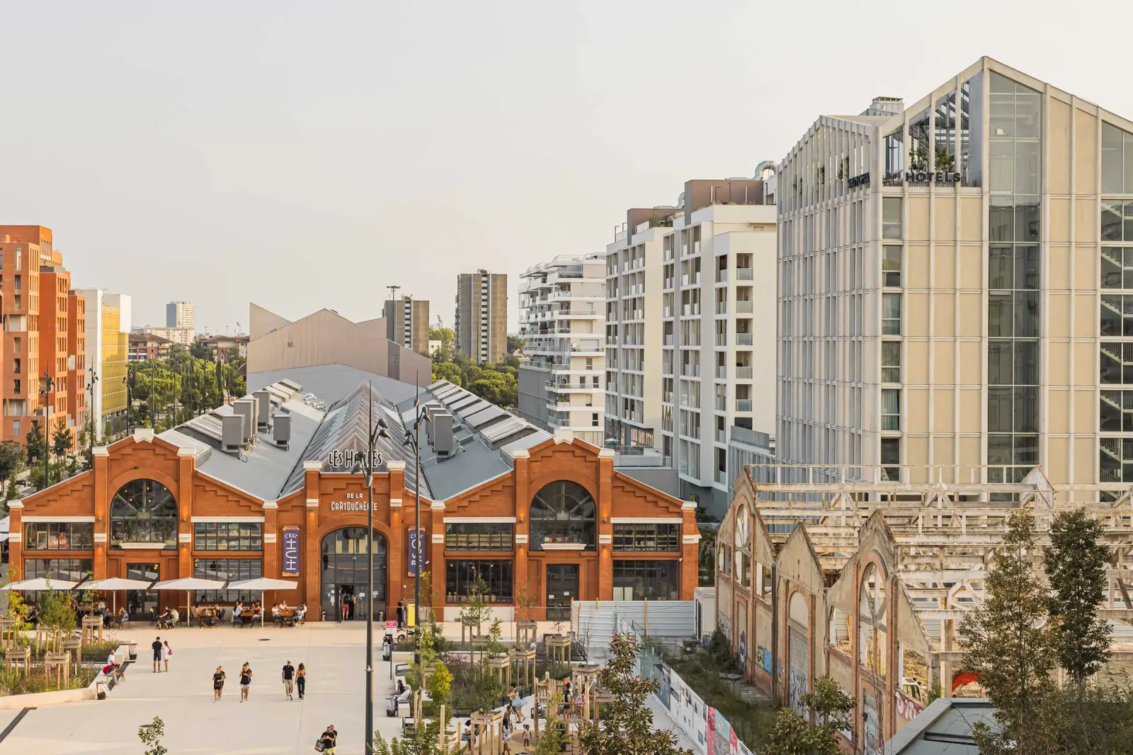 Wide view of the brick market hall with rooftop LES HALLES signage, an outdoor terrace and a public plaza with visitors at Les Halles de la Cartoucherie, Toulouse.