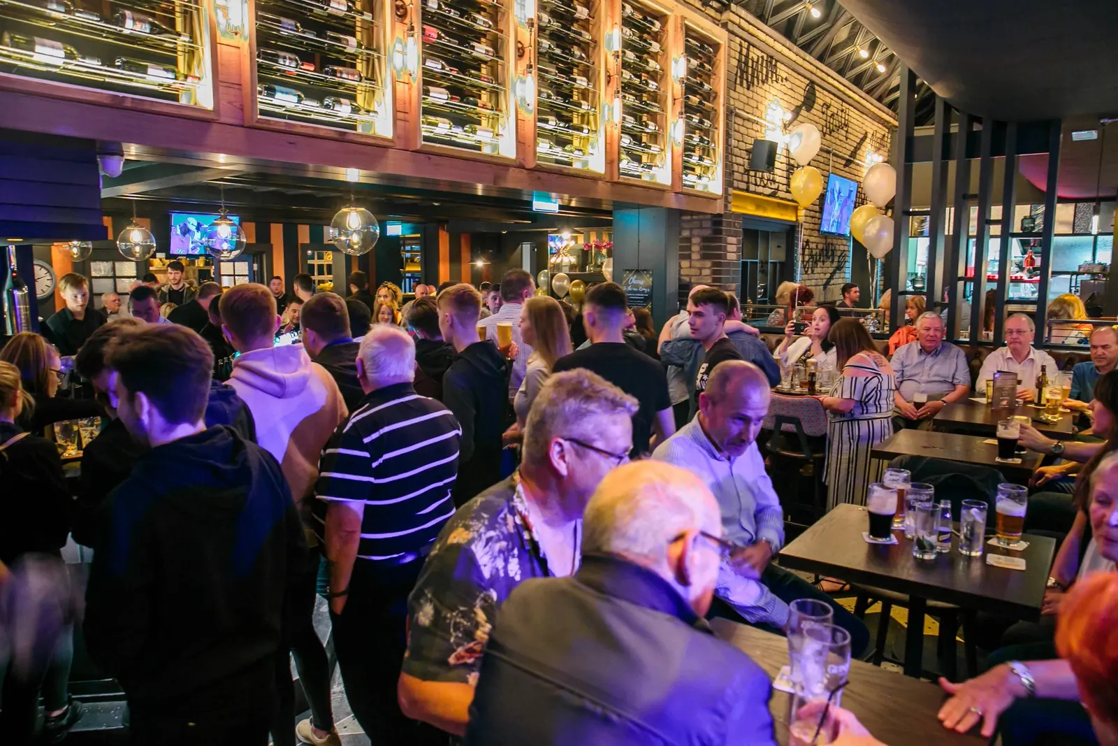Busy crowd with drinks under the lit wine wall at Kennys of Lucan, Dublin