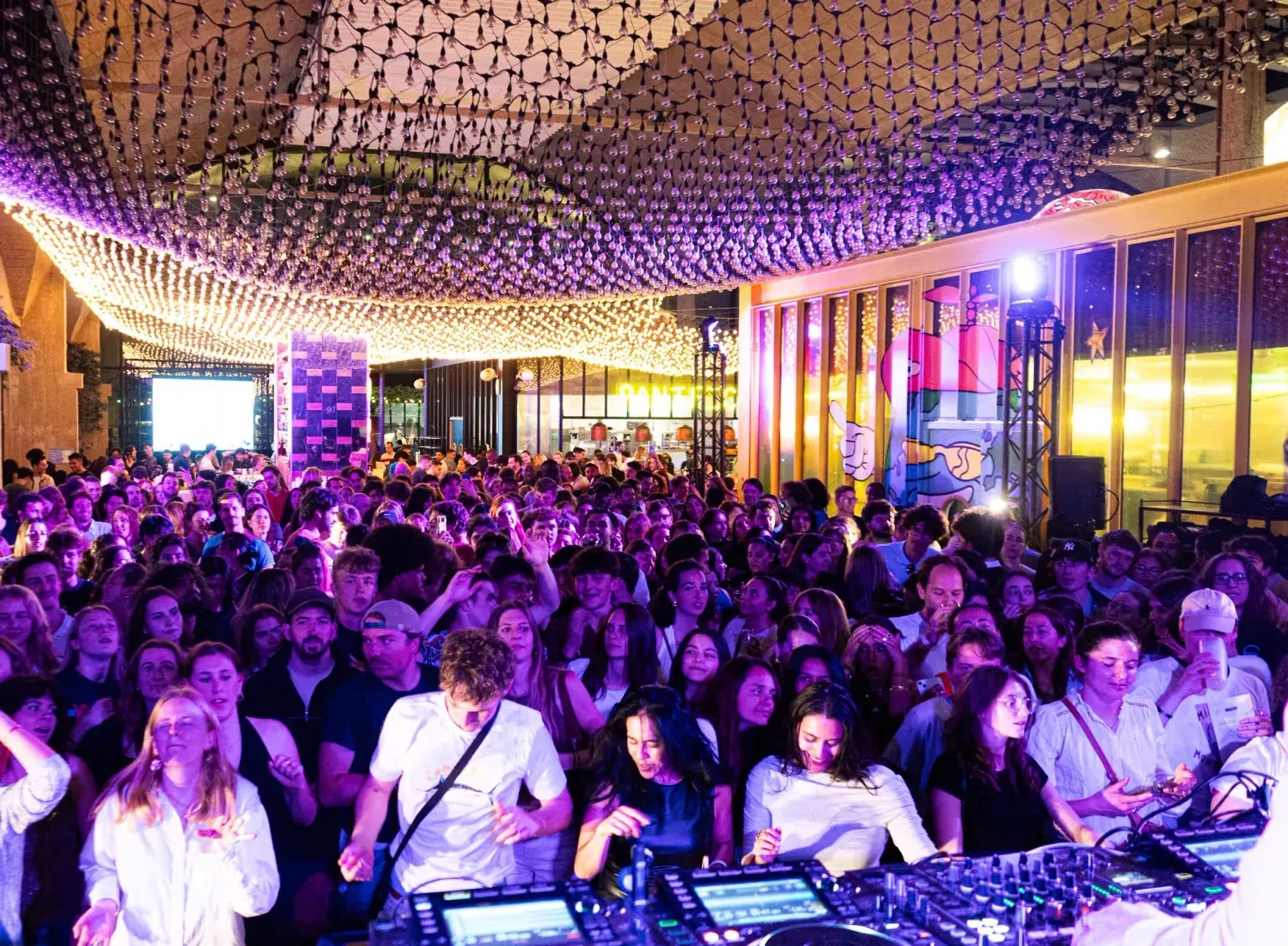 Nighttime crowd dancing under glowing string lights beside a DJ booth during a party, at La Felicità, Paris.