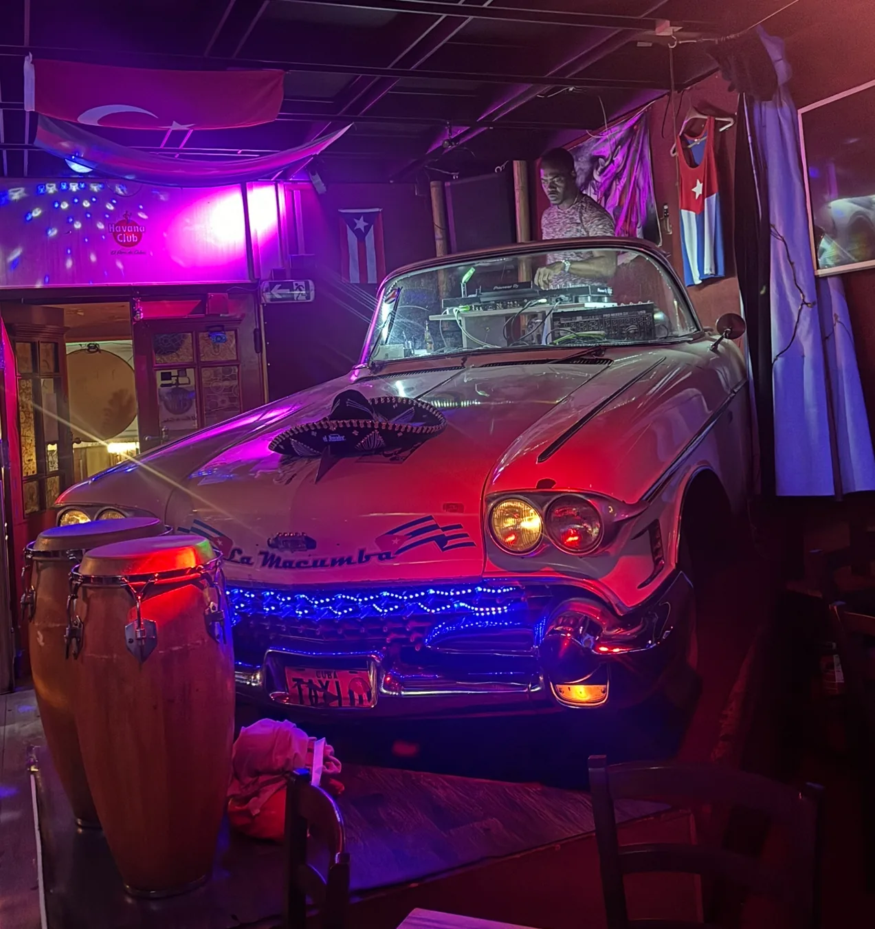 DJ booth behind a vintage car in the neon-lit interior of La Macumba Music Latino Bar, Cuban flags on the wall