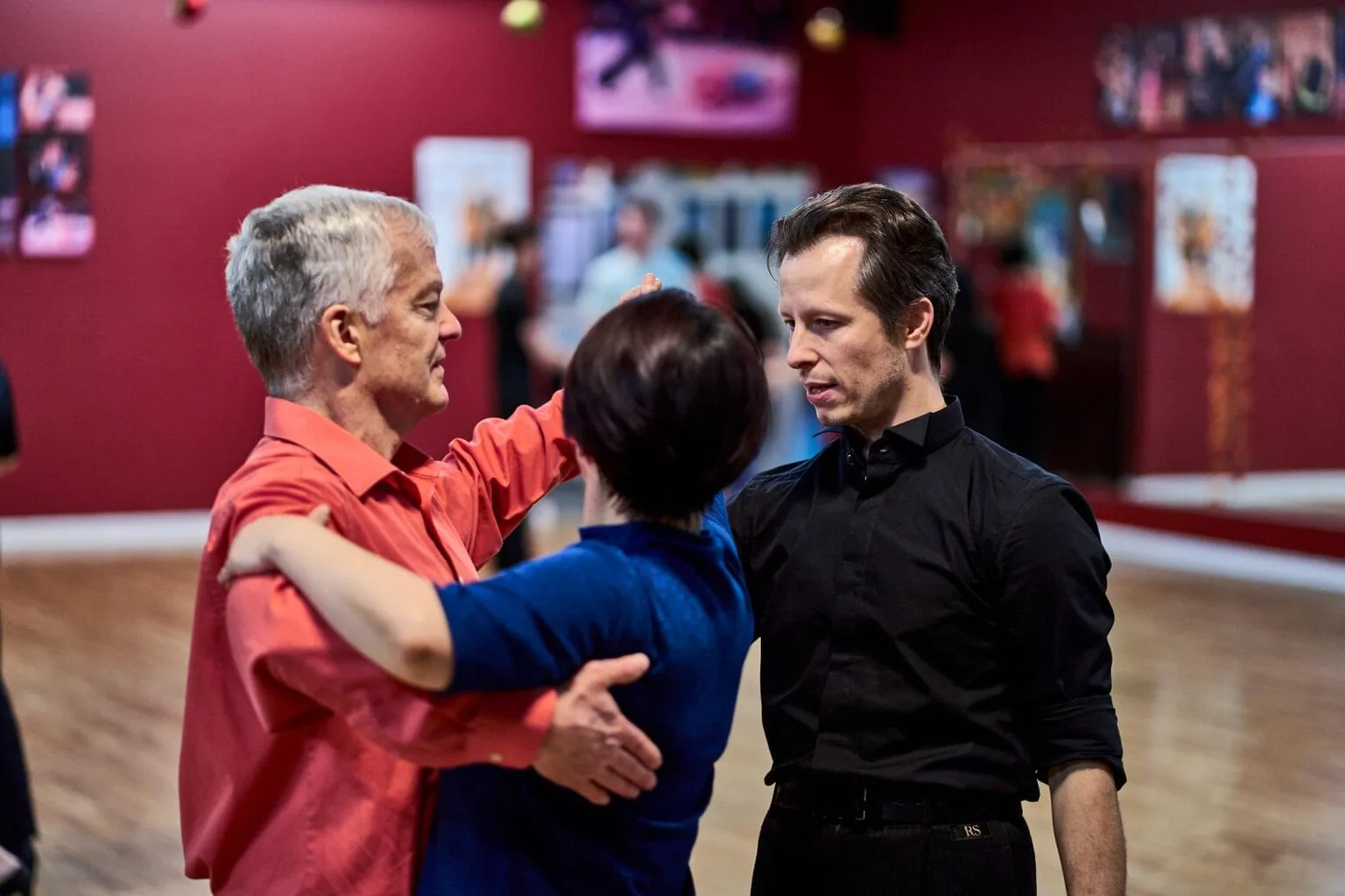 Instructor guiding a couple in a dance hold at Aria Ballroom, red walls with framed dance photos