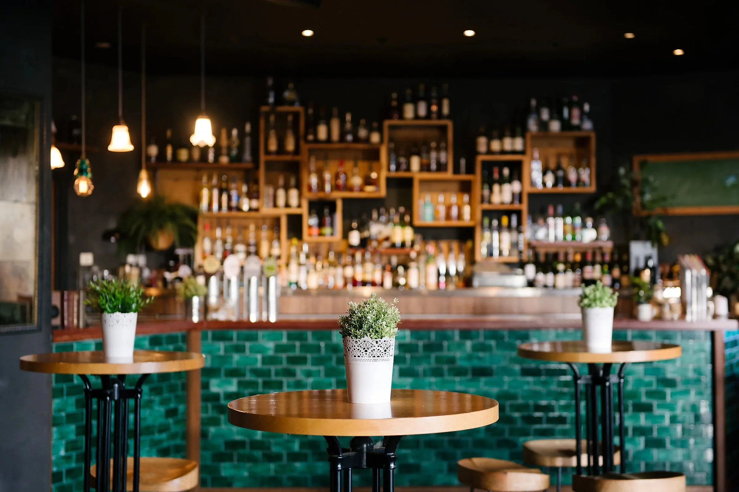 The main bar with its teal tiled counter, high round tables topped with potted plants and timber-crate shelving stocked with spirits at Loft West End, Brisbane.