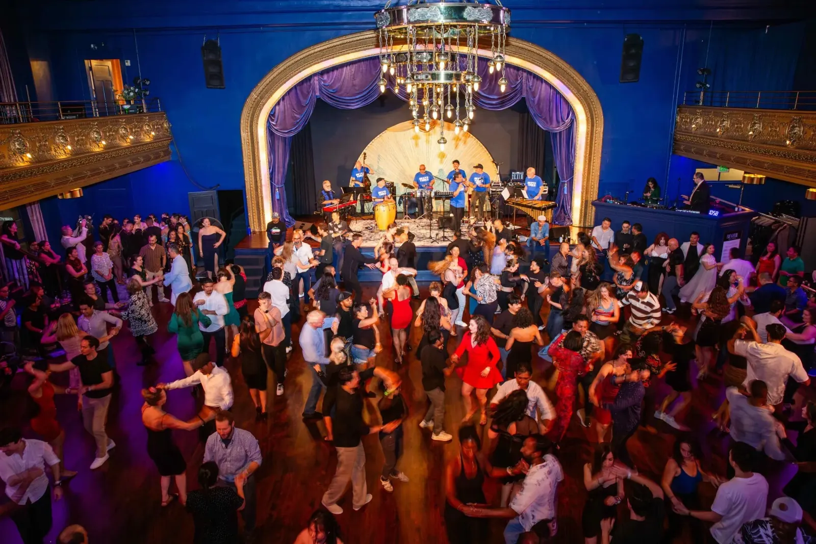 Packed social dance floor with couples dancing to a live Latin band on the ornate gold-framed stage, beneath a brass chandelier and deep blue balcony walls, at Reverie Ballroom, Seattle.