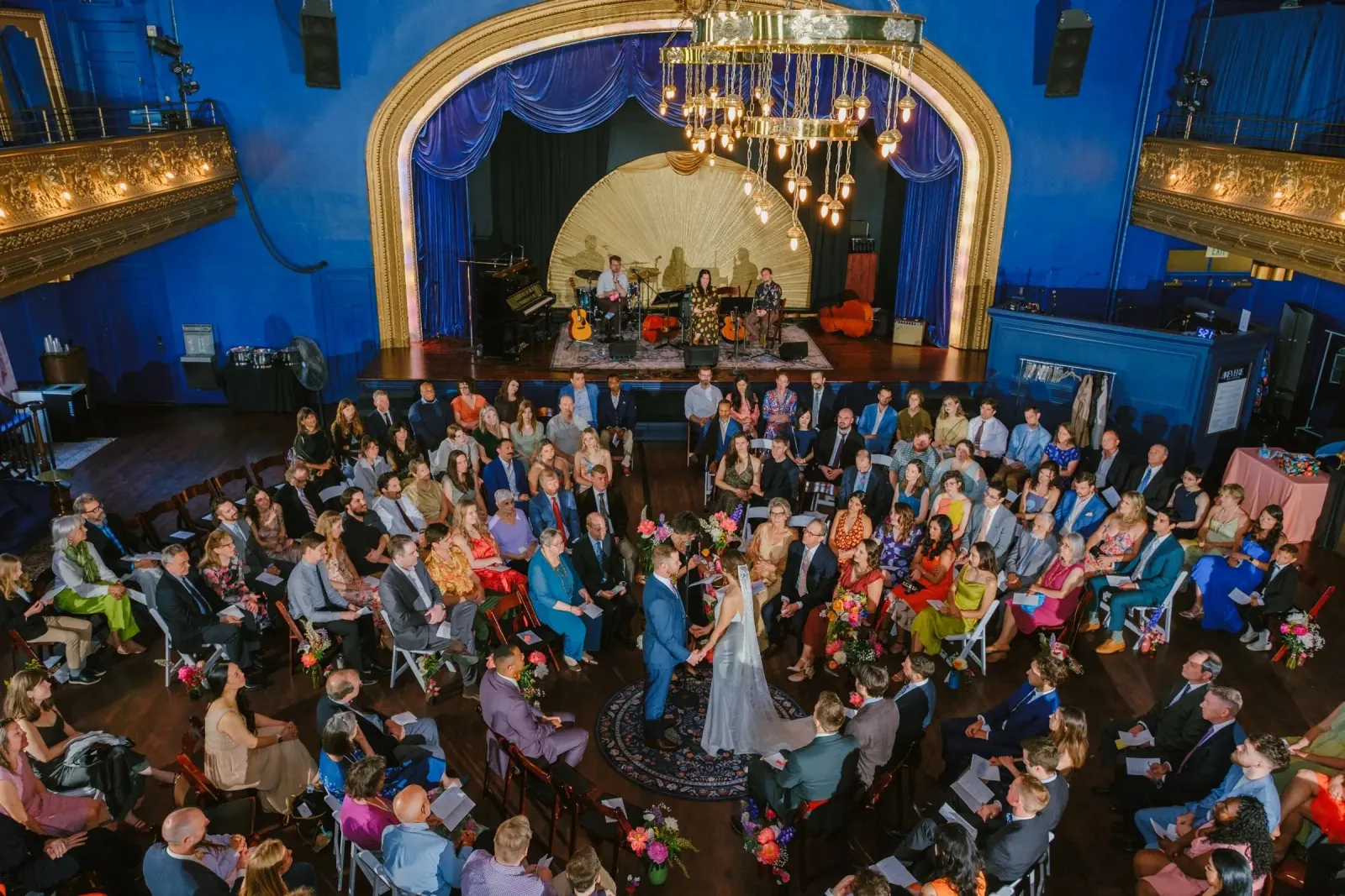 Grand ballroom interior with deep blue walls, a gilded proscenium arch, ornate balconies and a large brass chandelier, with guests seated around a central ceremony, at Reverie Ballroom, Seattle.