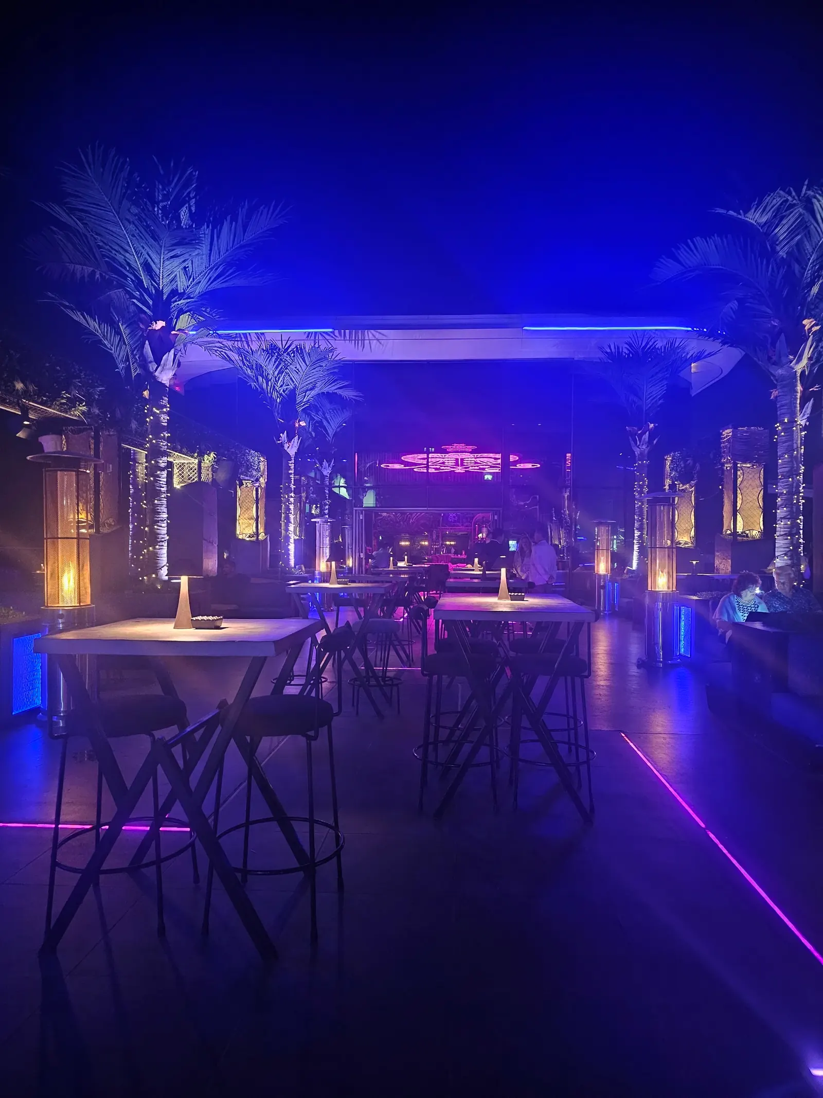 High tables and lit palms under blue and purple lighting at SoBe Rooftop Bar in Dubai at night