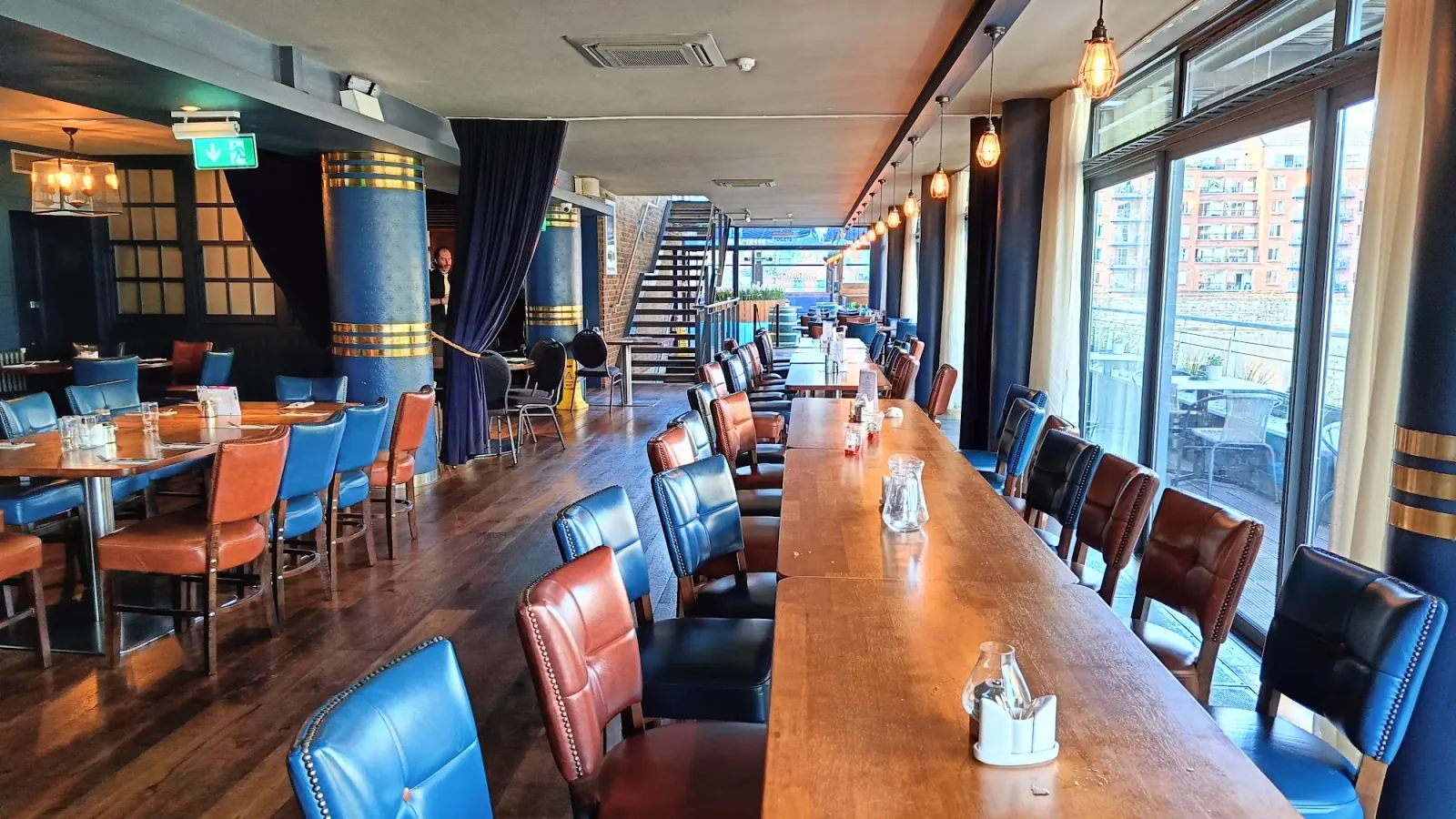 Wide interior of the Harbourmaster Bar in Dublin with a long communal table, blue and red leather chairs and waterfront-facing windows