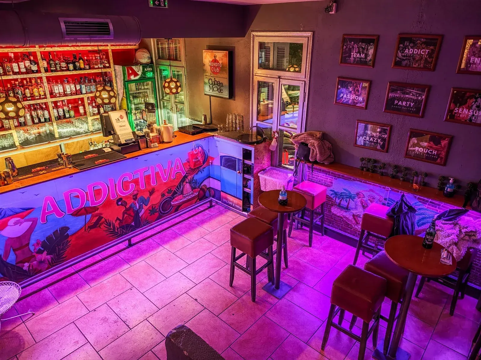 Pink and purple lit interior showing the bar with backlit spirit shelves, a hand-painted ADDICTIVA mural along the counter front, wooden bar stools and small round tables, and framed team photos on the wall, Addictiva Athens Bar, Athens