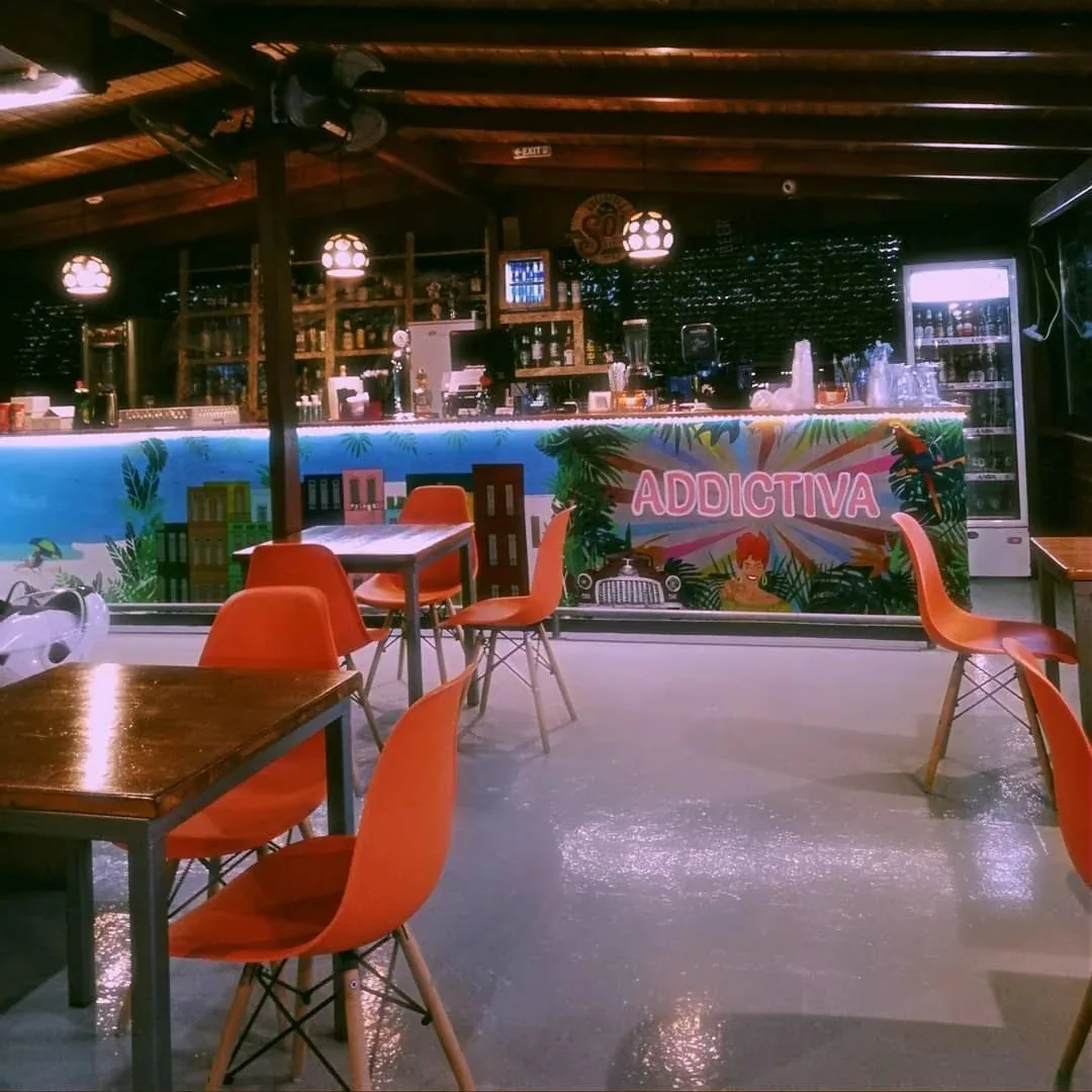 Covered open-air seating area with a wooden beamed ceiling, orange moulded chairs at wooden tables, and a tropical-themed bar counter with an ADDICTIVA mural and stocked drink fridges, Addictiva Athens Bar, Athens
