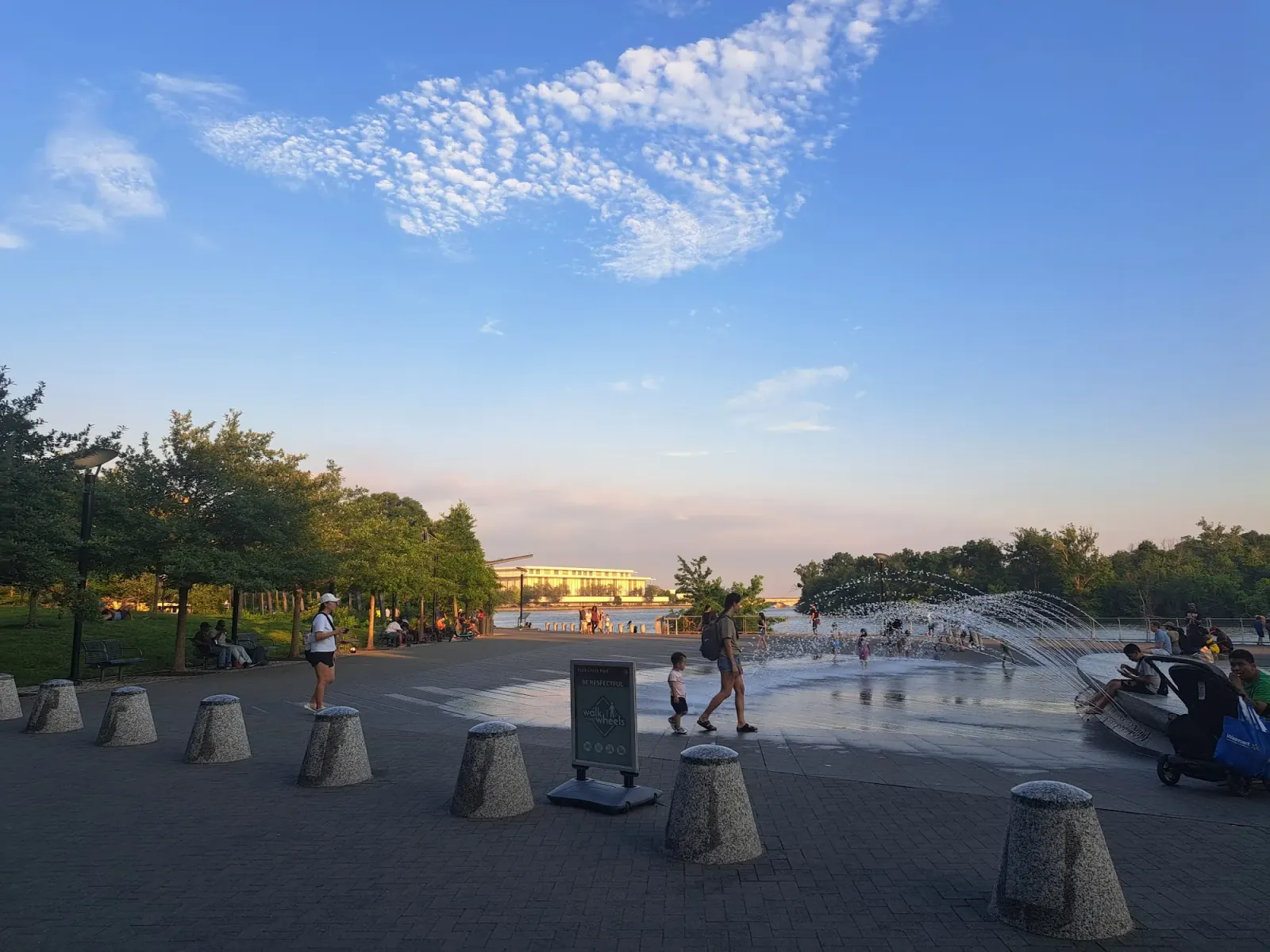 People gathering by the fountain plaza at Georgetown Waterfront Park with Kennedy Center across the Potomac