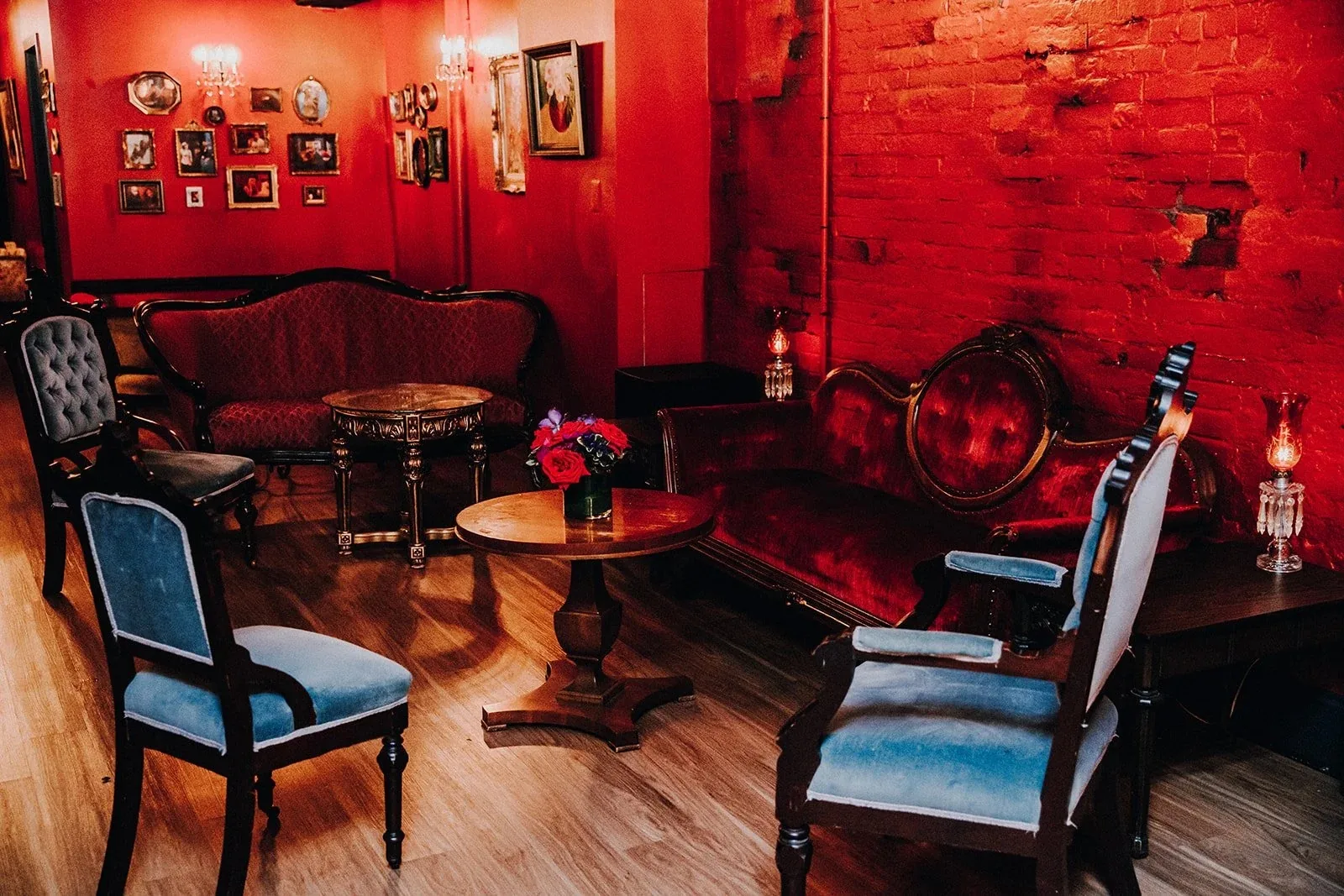 Dimly lit lounge with red walls, exposed brick, red velvet Victorian sofas and pale blue upholstered chairs around vintage wooden tables at Eighteenth Street Lounge, Washington.