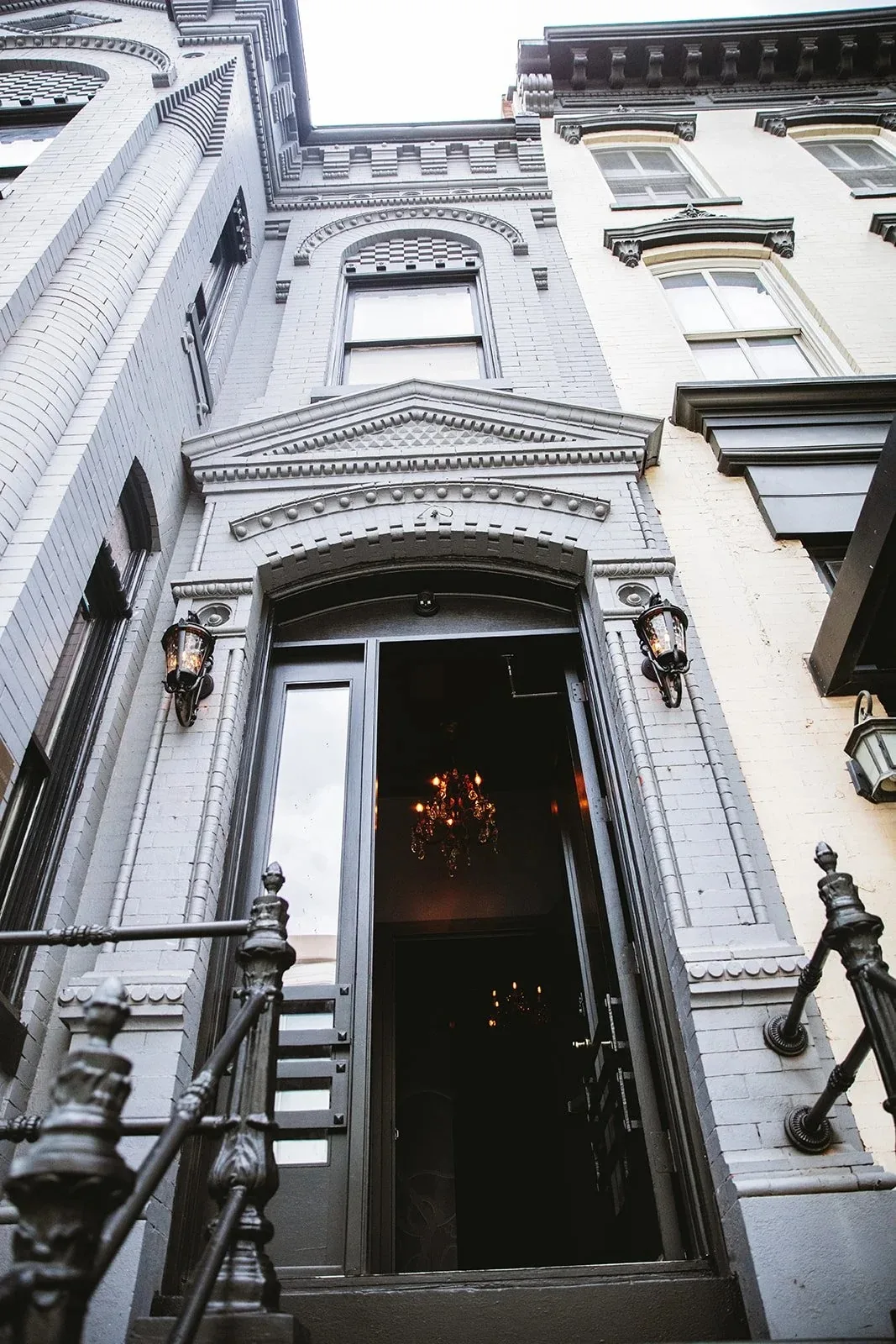 Low-angle view of the gray brick facade and entrance doorway flanked by lanterns, a chandelier glowing inside, at Eighteenth Street Lounge, Washington.